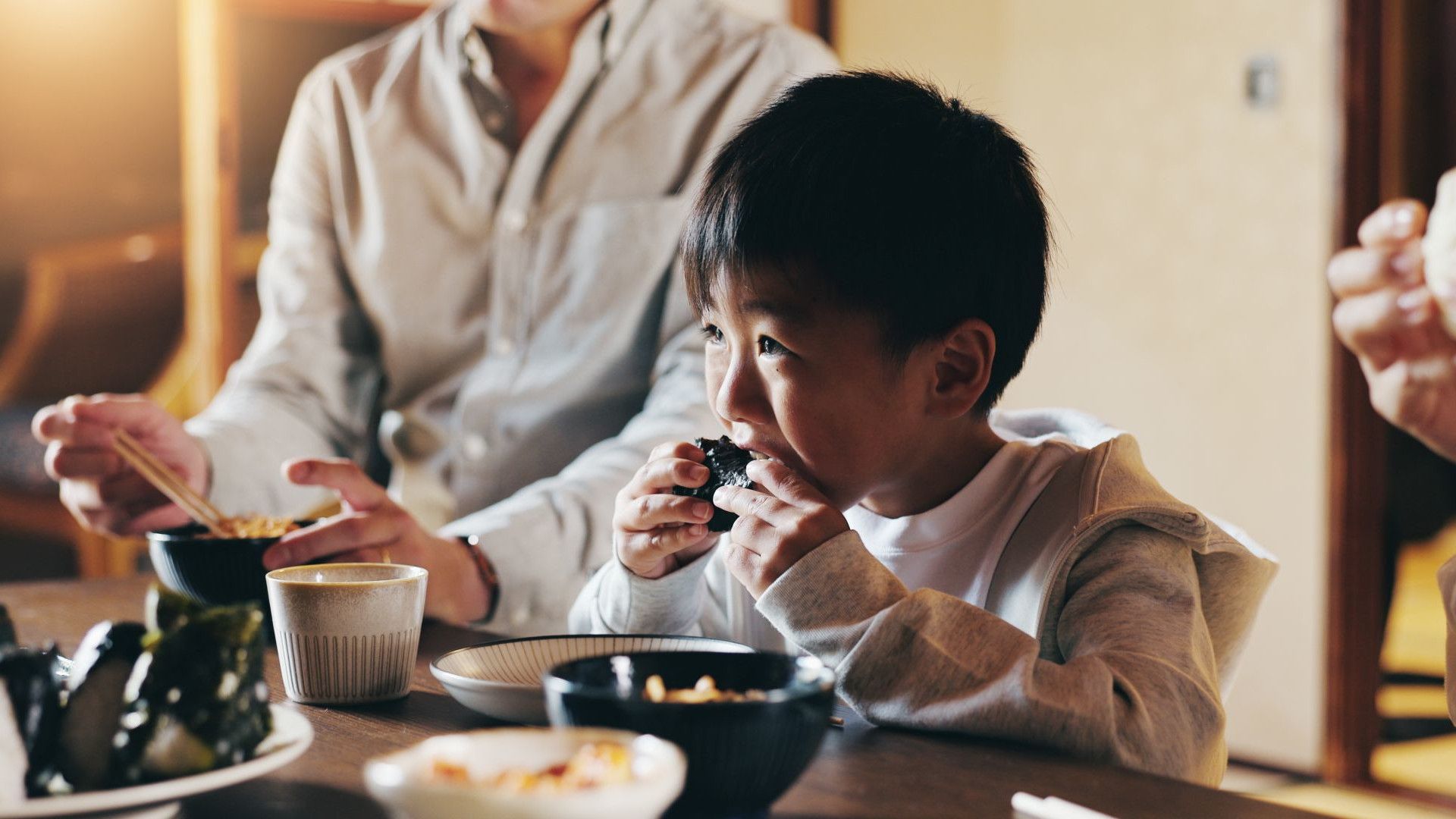 一個小孩和一個大人一起在木桌旁吃飯，桌上有碗、杯子和用海苔包裹的米飯。